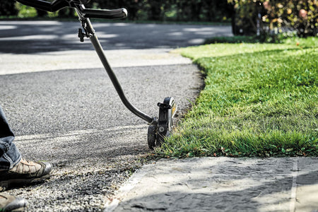 A landscaper using the DEWALT DWOAS4ED Edger Attachment to groom a grass border along an asphalt driveway, demonstrating the ease of movement provided by the large guide wheel.