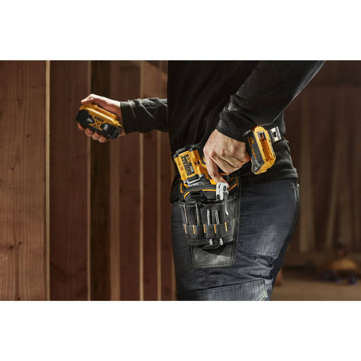A close-up action view of a professional user reaching for a cordless drill seated in the DEWALT ballistic nylon holster. The shot highlights the accessible placement of the 13 tool sleeves and the industrial durability of the fabric while the user navigates a construction workspace.
