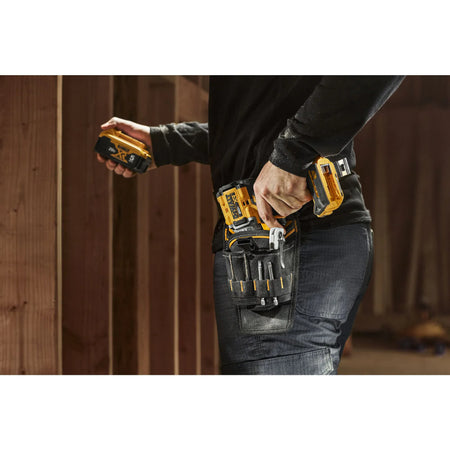 A close-up action view of a professional user reaching for a cordless drill seated in the DEWALT ballistic nylon holster. The shot highlights the accessible placement of the 13 tool sleeves and the industrial durability of the fabric while the user navigates a construction workspace.