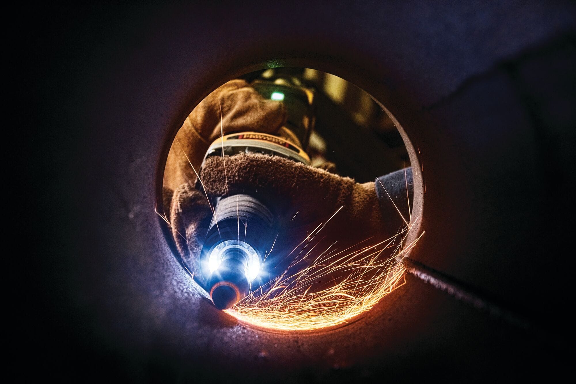 Industrial worker using a DEWALT DWE4997VS die grinder inside a metal pipe, wearing protective gloves as bright sparks fly during precision grinding and finishing work.
