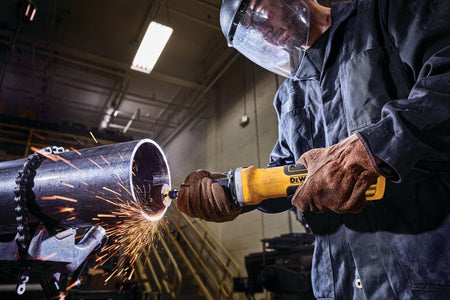 Operator wearing full protective gear using a DEWALT DWE4997VS die grinder on the edge of a large metal pipe, producing bright sparks during heavy‑duty grinding and fabrication work.