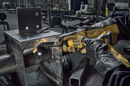 Professional metalworker wearing heavy leather gloves using the DEWALT DCG432XGE2 20V MAX 4 inch extended cut-off tool to slice through a steel pipe, demonstrating narrow space cutting capabilities with flying sparks.