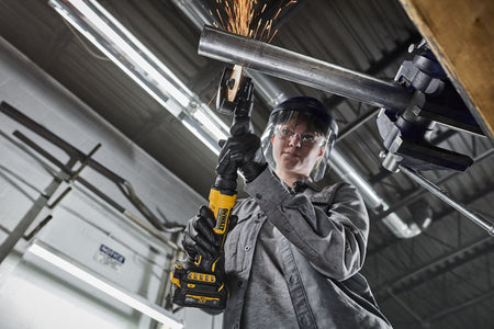 Metalworker wearing safety gear performing an overhead cut on a suspended steel pipe using the DEWALT DCG432XGE2 4 inch cordless extended cut-off tool, illustrating ergonomic control and cordless convenience.