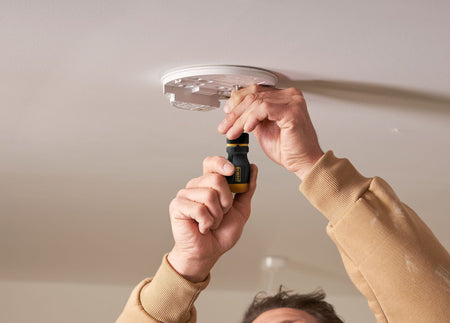 Contractor using the DEWALT stubby ratcheting screwdriver to install a ceiling-mounted smoke detector, showcasing the tool's effectiveness for overhead work in tight residential spaces.