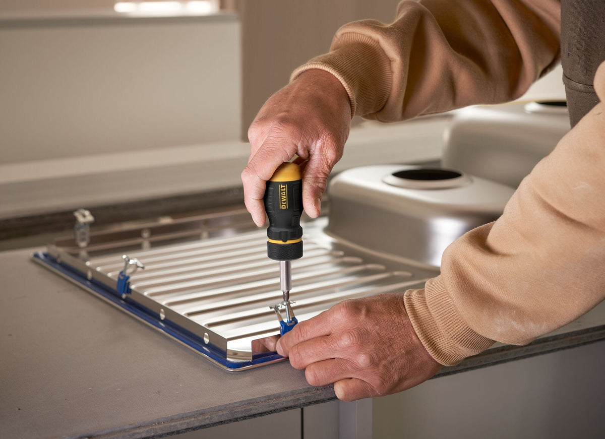 Action shot of a professional using the DEWALT stubby ratcheting screwdriver to secure fasteners in a cramped kitchen sink installation, demonstrating the tool's compact maneuverability.