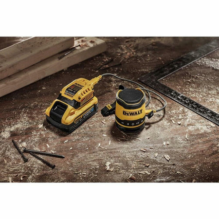 The DEWALT mini Bluetooth speaker on a dusty workbench, being charged by a USB cable connected to a DEWALT 20V MAX USB Power Source adapter.