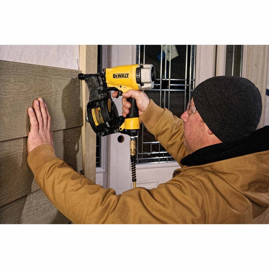 Person using a DEWALT pneumatic nail gun to install textured siding on a wall, with one hand pressing the material and the other operating the tool connected to an air hose.
