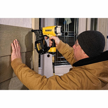 Person using a DEWALT pneumatic nail gun to install textured siding on a wall, with one hand pressing the material and the other operating the tool connected to an air hose.