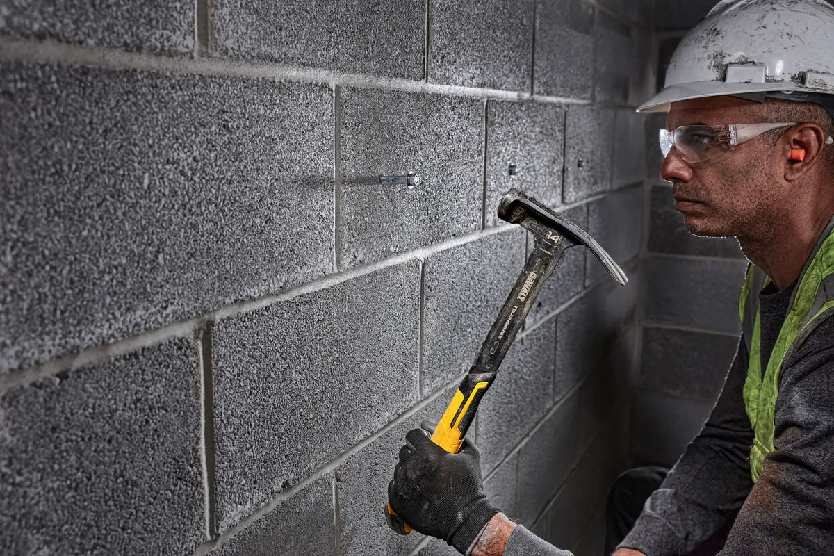 A worker holding the DEWALT 14 oz steel framing hammer while inspecting a concrete masonry unit wall, demonstrating the tool's balanced weight distribution for reduced fatigue.
