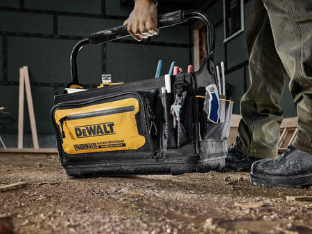 Lifestyle image of a person holding a DEWALT tool tote filled with hand tools in a construction setting. The black and yellow tote displays “WATER RESISTANT CONSTRUCTION” text and organized exterior pockets. The person stands on a dusty floor with wood and building materials in the background
