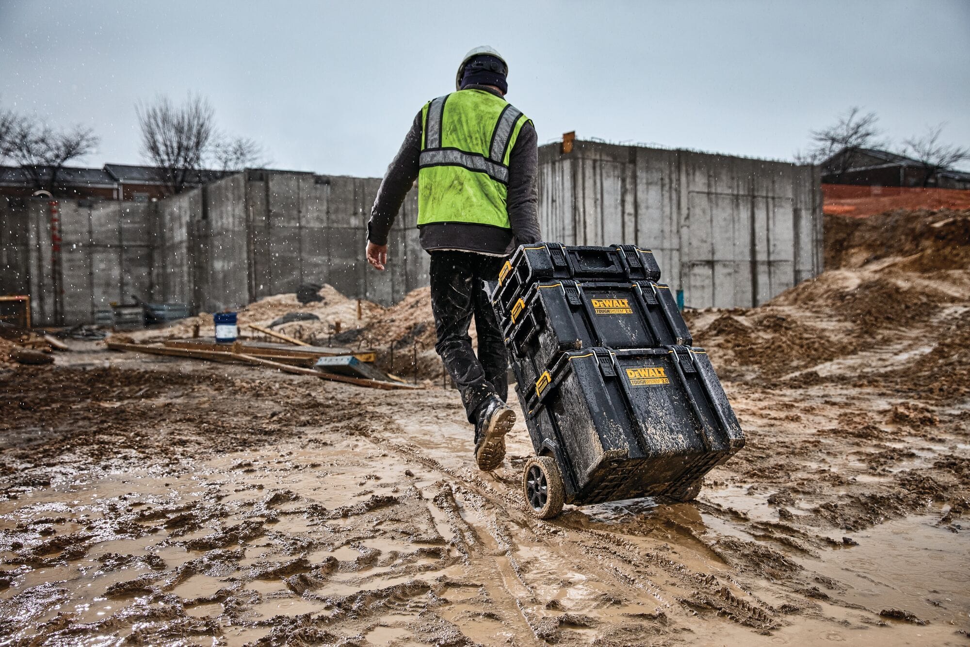 Worker pulling stacked DEWALT ToughSystem tool boxes across wet muddy construction site demonstrating weather resistant durability and mobile tool transport in harsh jobsite conditions.
