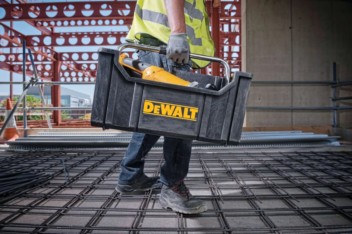 An action lifestyle shot of a professional contractor carrying the DEWALT ToughSystem tote on a construction jobsite. The tote is filled with a power tool, illustrating the convenience of the open-top design for quick access to frequently used equipment while navigating a work area.