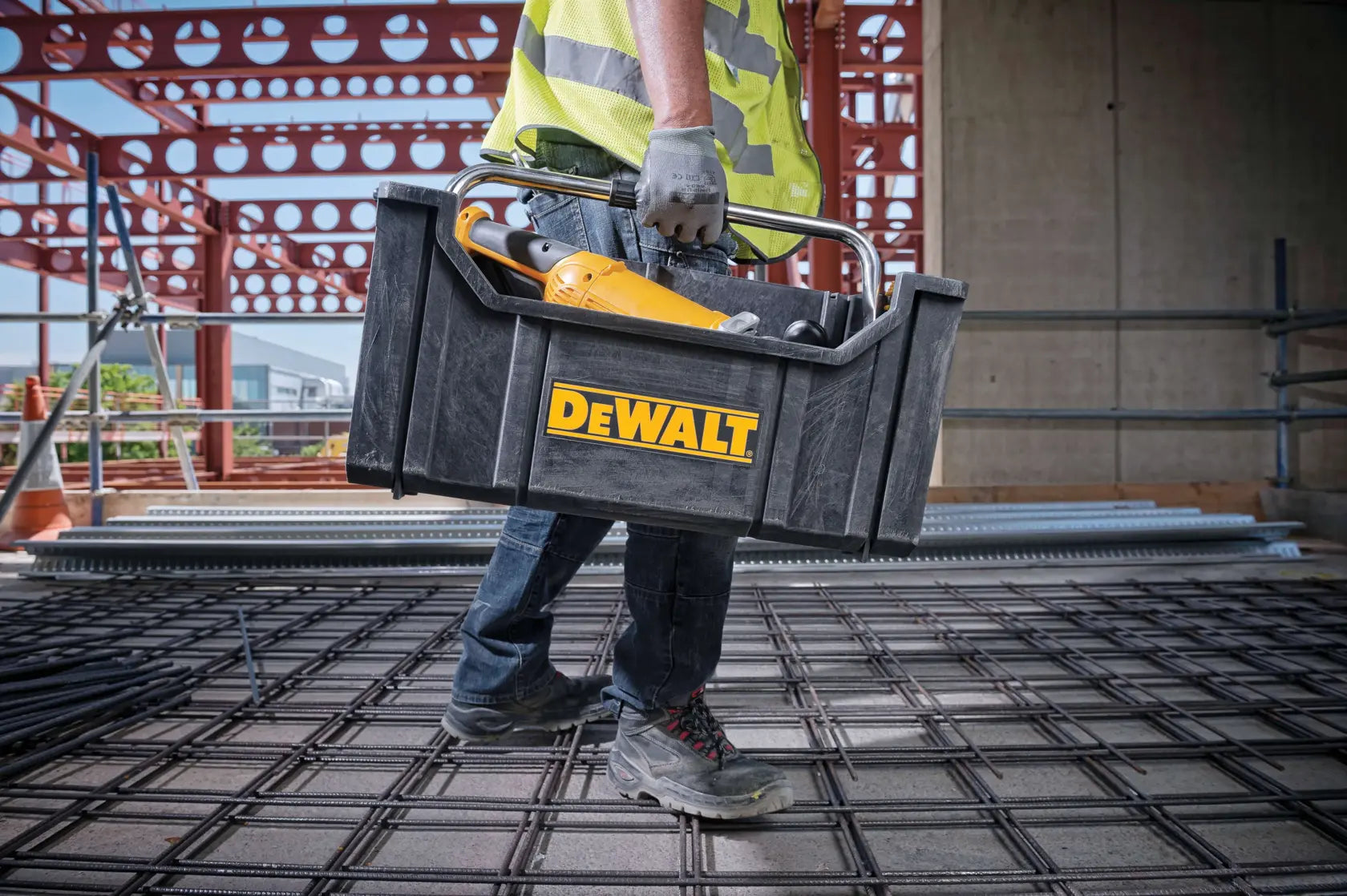 An action lifestyle shot of a professional contractor carrying the DEWALT ToughSystem tote on a construction jobsite. The tote is filled with a power tool, illustrating the convenience of the open-top design for quick access to frequently used equipment while navigating a work area.