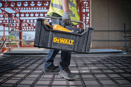 An action lifestyle shot of a professional contractor carrying the DEWALT ToughSystem tote on a construction jobsite. The tote is filled with a power tool, illustrating the convenience of the open-top design for quick access to frequently used equipment while navigating a work area.