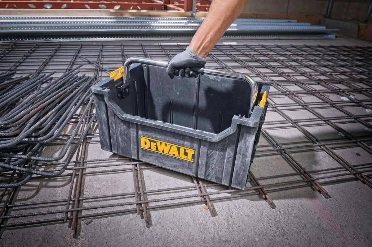 A high-angle action shot of a worker’s hand grabbing the metal handle of the DEWALT tool tote. The container is sitting on a steel-reinforced floor, highlighting the industrial-grade durability and portability of the bulk storage container for professional tradespeople.