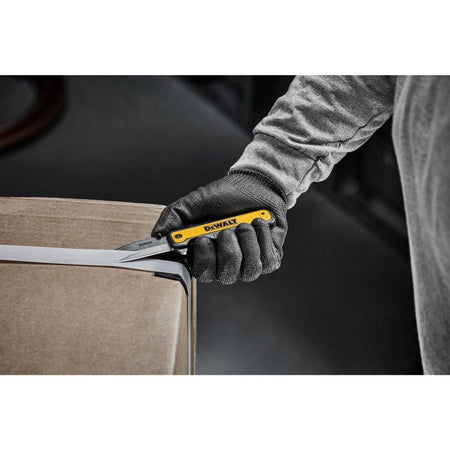 Person wearing gloves using a DEWALT DWHT10993 pocket knife to cut a white plastic strap on a cardboard box in an industrial setting.