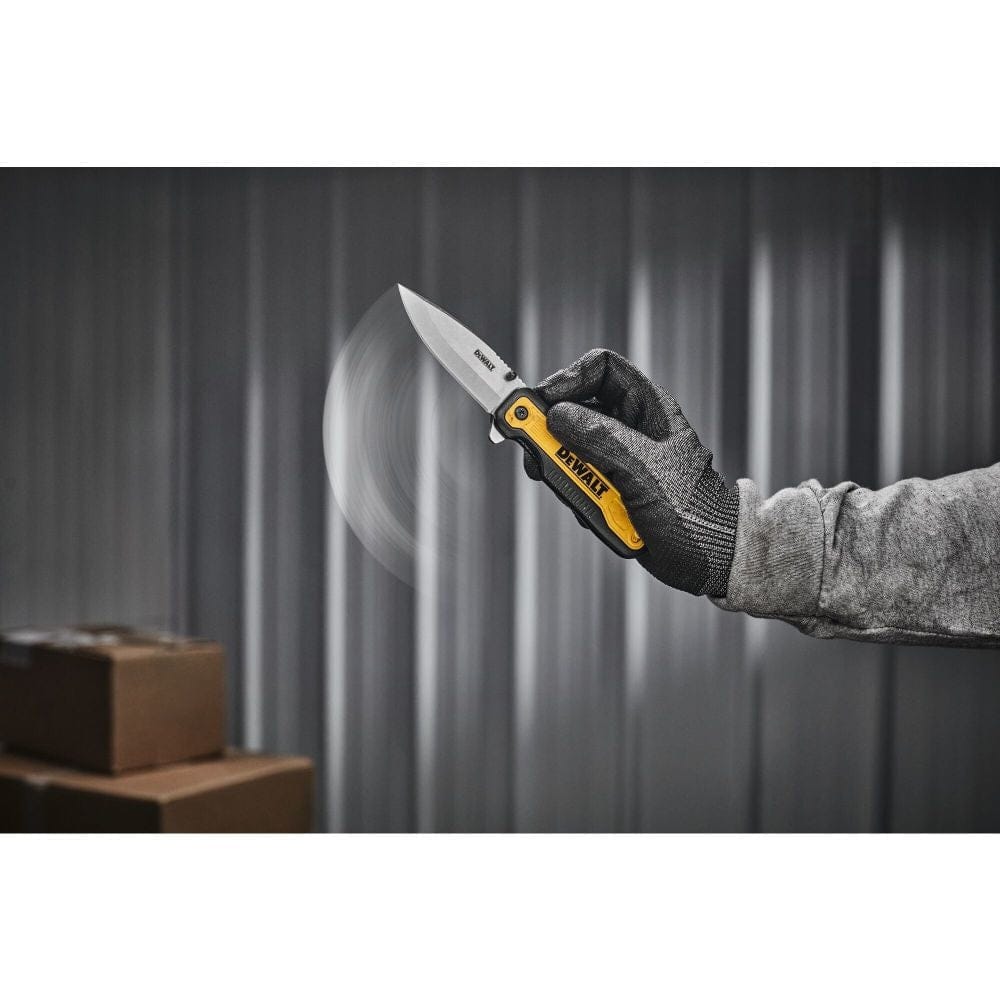 Person wearing gloves flipping open a DEWALT DWHT10993 folding knife with motion blur; background includes corrugated metal wall and cardboard boxes.