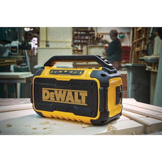 DeWalt Bluetooth speaker sits on a wooden workbench in a workshop. The rugged yellow and black speaker features a top handle and front control panel, surrounded by tools and shelving.