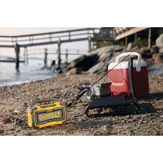 DeWalt Bluetooth speaker placed on a sandy beach near a cooler, fishing rod, and tackle box. The speaker provides portable audio during a recreational outing by the water.
