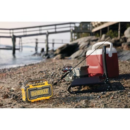 DeWalt Bluetooth speaker placed on a sandy beach near a cooler, fishing rod, and tackle box. The speaker provides portable audio during a recreational outing by the water.
