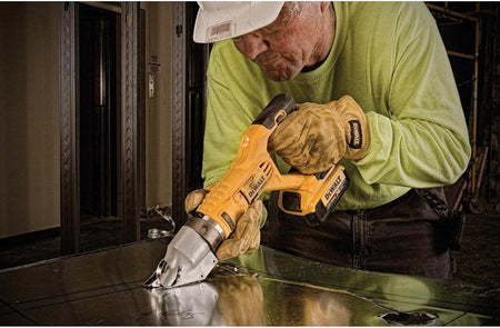 Person wearing safety gear using a DEWALT electric shear to cut sheet metal in a construction setting, demonstrating tool application in industrial environments.