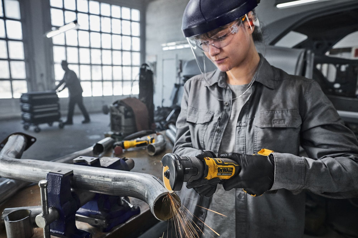 Automotive workshop application graphic showing a technician wearing a clear face shield and black nitrile safety gloves utilizing the DeWalt 20V MAX compact cut-off saw to modify a bent steel exhaust line pipe secured inside a heavy bench vise, throwing down a stream of sparks