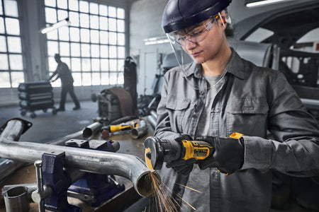 Automotive workshop application graphic showing a technician wearing a clear face shield and black nitrile safety gloves utilizing the DeWalt 20V MAX compact cut-off saw to modify a bent steel exhaust line pipe secured inside a heavy bench vise, throwing down a stream of sparks
