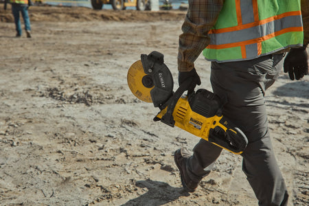 Worker carrying the portable DeWalt DCS692 60V cut-off saw by its front handle across an outdoor jobsite.