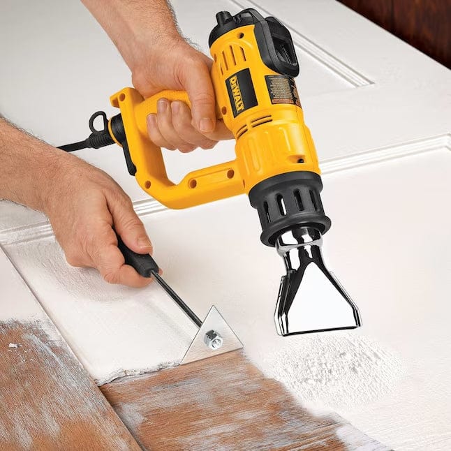 A user stripping white paint from a wooden surface using the heat gun to soften the material while scraping it away with a handheld metal triangle scraper.
