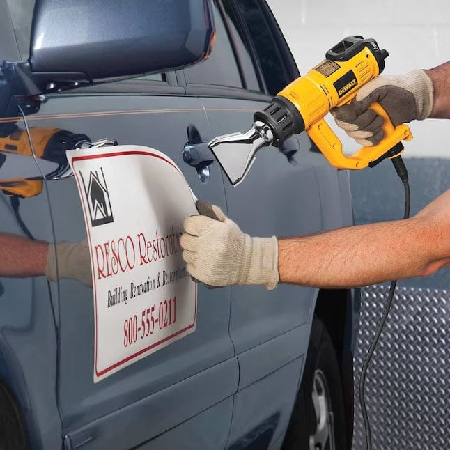 A worker using the DeWalt heat gun equipped with a wide fishtail nozzle to heat and peel a large decal off the side of a vehicle door.