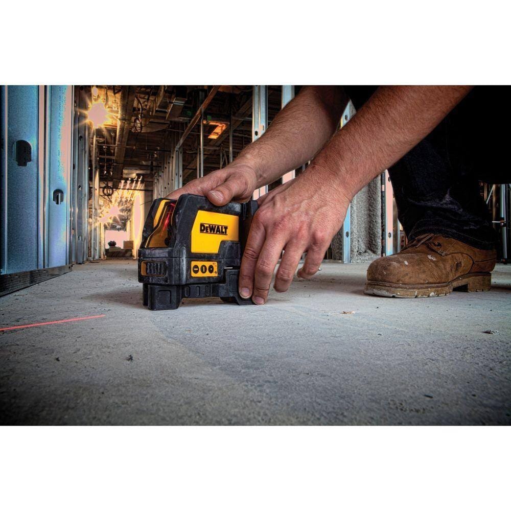 An action photograph of a professional worker in boots and work pants crouching on a concrete floor to position the DeWalt DW0822 laser level, which is projecting a red alignment line across the floor in a commercial construction setting.