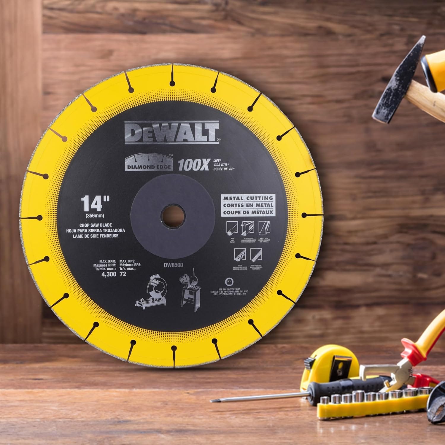 A lifestyle product shot of the DeWalt 14-inch diamond edge metal cutting wheel resting upright on a rustic wood workbench, surrounded by blurred workspace hand tools including a hammer, pliers, sockets, and a tape measure under ambient lighting.
