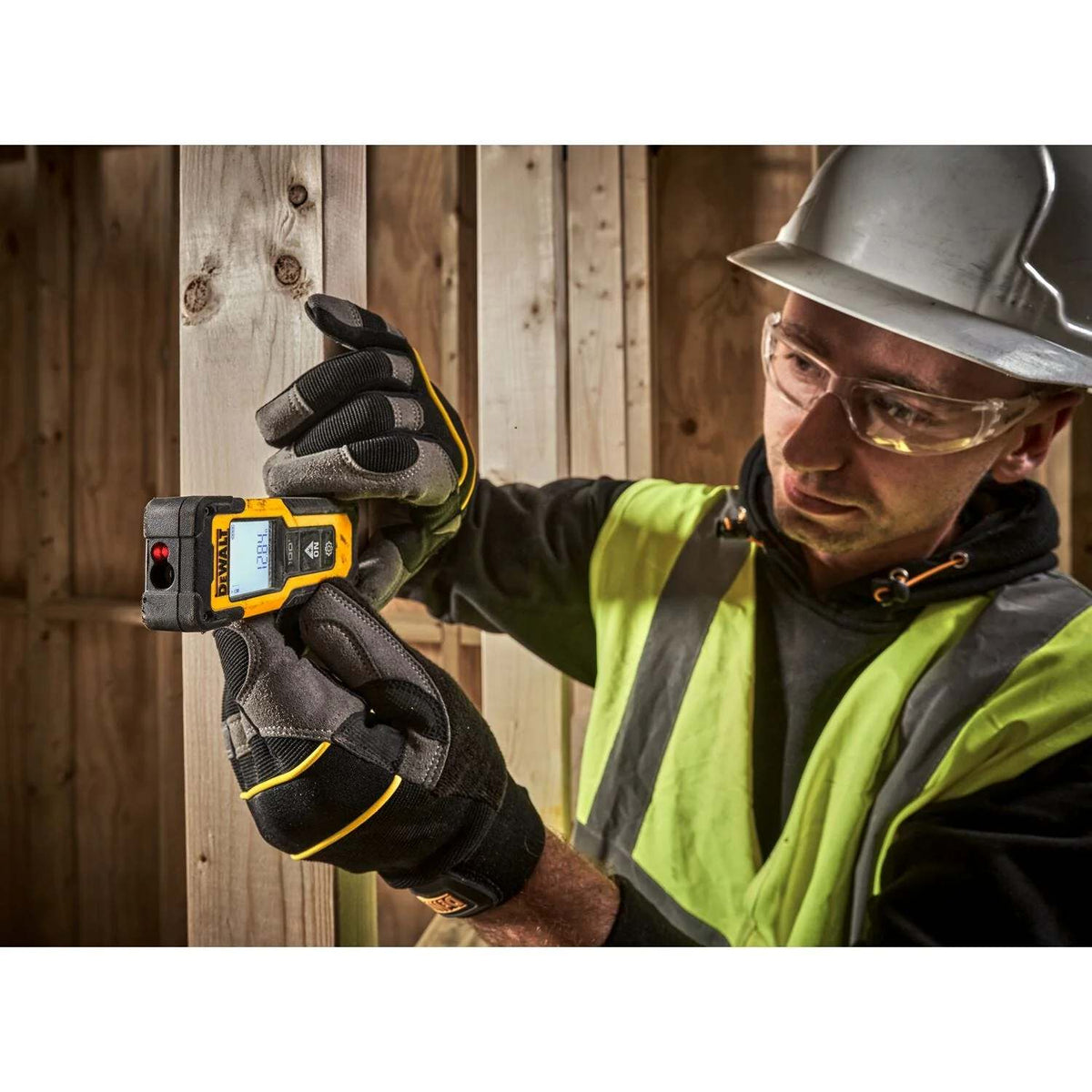 A construction worker wearing a hard hat and safety glasses holding the DeWalt laser measurer against a wooden stud to capture a distance reading.