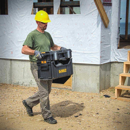 A worker walking across a dirt jobsite carrying the DeWalt tote by its side handles, demonstrating its stacking capability with a smaller ToughSystem organizer box latched securely on top.