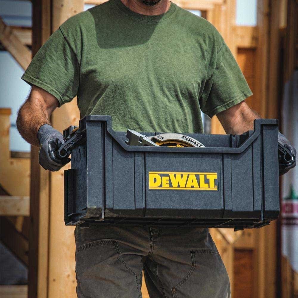 Construction worker holding the DeWalt open-top tote with both hands by the integrated side handles, transporting a circular saw placed inside the deep storage compartment.