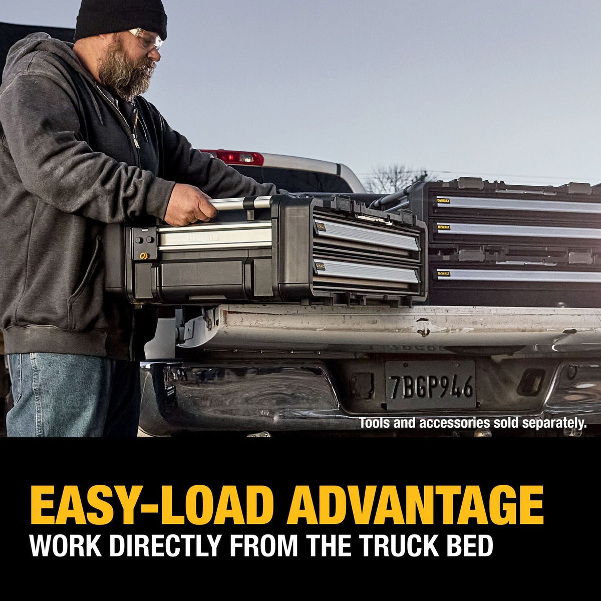 An outdoor jobsite action photo showing a beard contractor wearing a black winter beanie, safety glasses, and a grey hoodie lifting a DEWALT ToughSystem 2.0 drawer box unit directly out of the open tailgate bed of a silver pickup truck, highlighted by lower text stating EASY-LOAD ADVANTAGE WORK DIRECTLY FROM THE TRUCK BED.