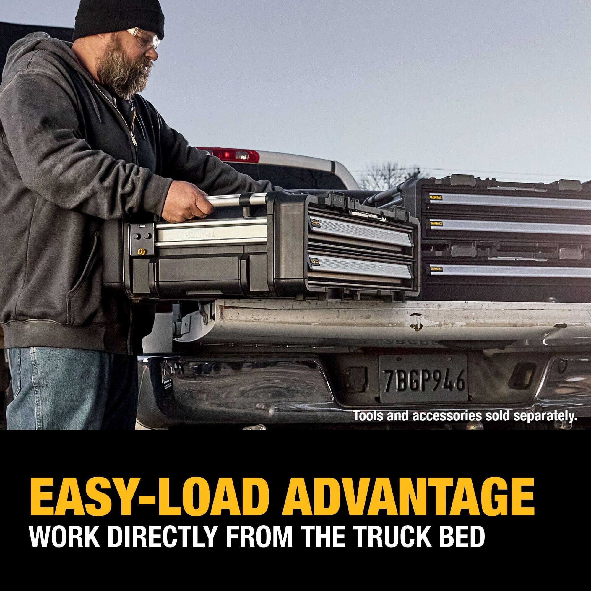 An outdoor jobsite action photo showing a beard contractor wearing a black winter beanie, safety glasses, and a grey hoodie lifting a DEWALT ToughSystem 2.0 drawer box unit directly out of the open tailgate bed of a silver pickup truck, highlighted by lower text stating EASY-LOAD ADVANTAGE WORK DIRECTLY FROM THE TRUCK BED.
