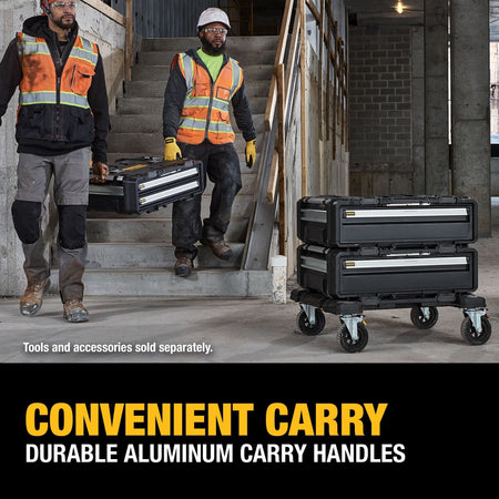 An indoor jobsite application lifestyle photo of two construction tradesmen wearing white hard hats and safety vests carrying a DEWALT modular drawer box unit down a concrete staircase at a commercial construction building site, while a separate stacked cart assembly stands on the floor, complete with lower text reading CONVENIENT CARRY DURABLE ALUMINUM CARRY HANDLES.
