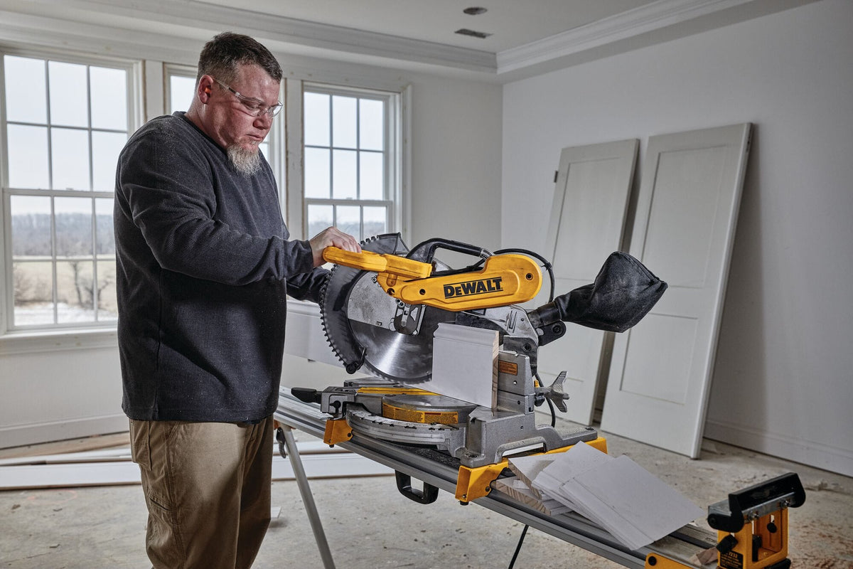 Trim carpenter cutting white baseboard molding held vertically against the sliding fence of the DeWalt DWS716 miter saw on a jobsite stand.