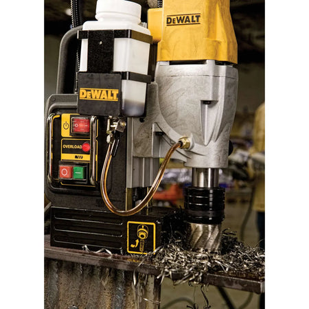 A jobsite macro photograph of the Dewalt 2-speed magnetic drill press in operation, showing an annular cutter boring directly into a thick structural steel beam surrounded by metal shavings, with the fluid coolant bottle mounted to the side of the housing.