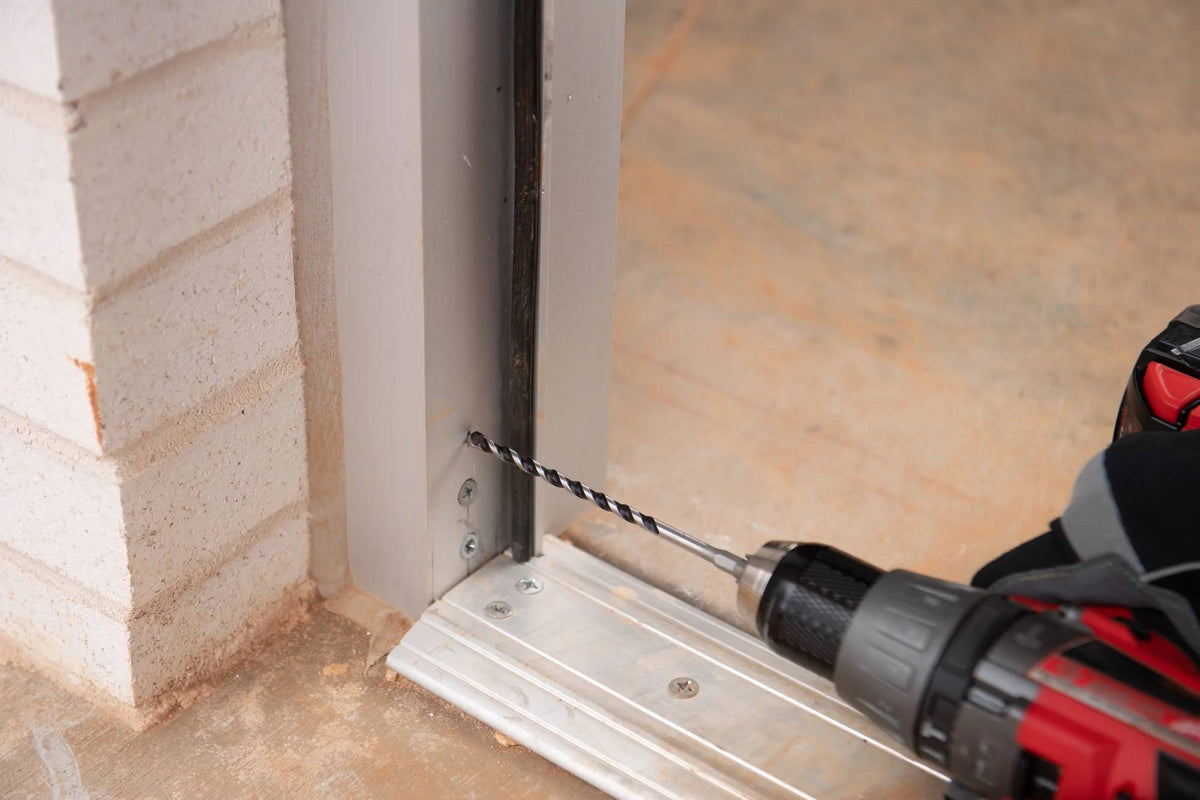 Person using a power drill to bore into a metal door frame near a concrete floor, demonstrating the drill bit’s effectiveness in metal and masonry applications