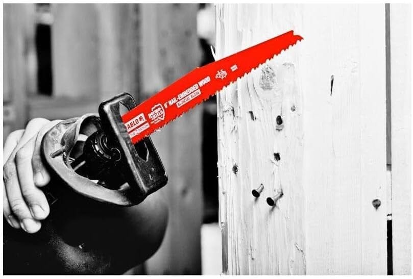 A black and white action photo with a color-highlighted red Diablo blade being used in a reciprocating saw to cut through a piece of wood with several nails embedded in it.