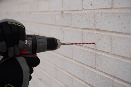 Action shot of a Diablo Red Granite hammer drill bit attached to a cordless drill while boring a precise hole into a white brick masonry wall.