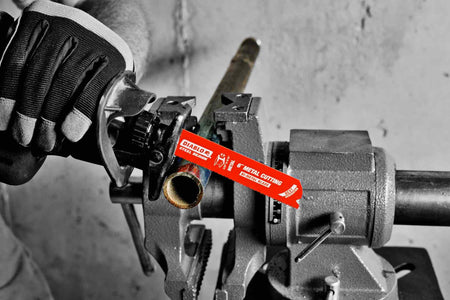 A selective-color action shot of a reciprocating saw, held by a gloved hand, using a red Diablo metal cutting blade to cut a pipe held in a bench vise.