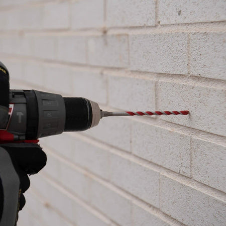 Close-up of a person using a cordless drill with a red Diablo bit to drill into a white brick wall, demonstrating masonry application and tool compatibility.