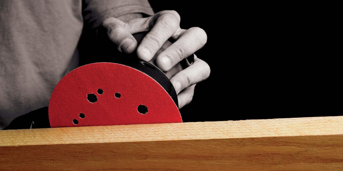 Action shot of a professional using a random orbital sander equipped with a Diablo red ceramic sanding disc to smooth the edge of a light-colored wooden board.