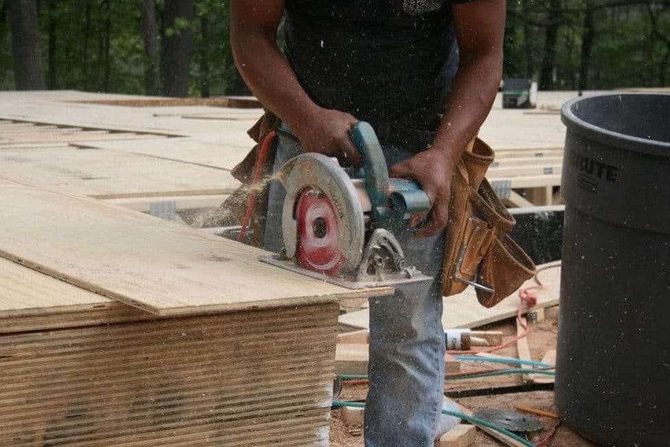 Person using circular saw to cut plywood on construction site; sawdust flying and tool belt visible.