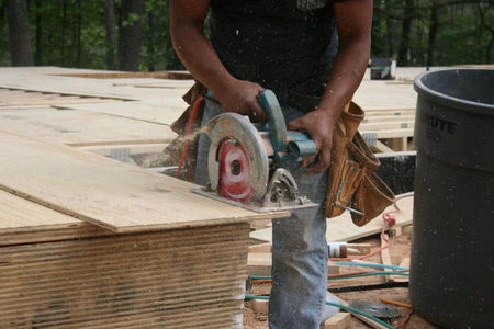 Person using circular saw to cut plywood on construction site; sawdust flying and tool belt visible.
