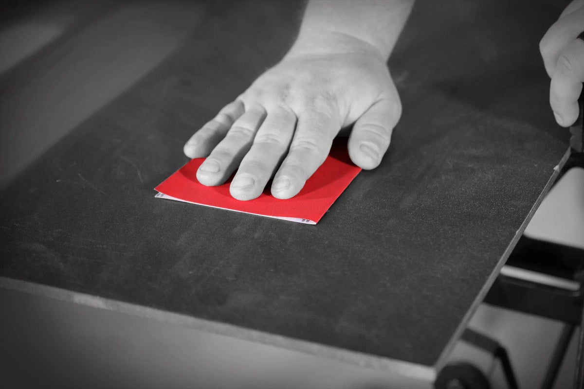 A hand using a piece of the red 60-grit sandpaper to sand a dark surface in a workshop setting.