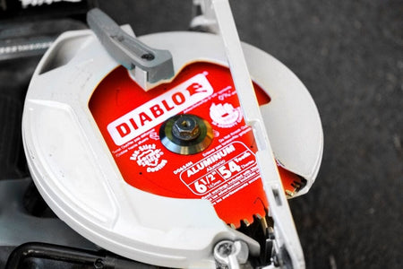 A close-up action photograph of the Diablo D0654NA 54-tooth blade installed in a cordless circular saw. The image shows the red blade through the saw's clear guard, demonstrating its compatibility with modern battery-powered cutting tools for non-ferrous metal applications.
